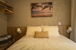Une chambre d’hôtel avec un lit, deux oreillers, une lampe fixée au mur, une étagère avec un sac et un tableau représentant une scène de plage.