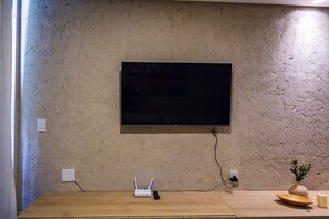 Une pièce avec un téléviseur fixé au mur, un mur à texture, une table en bois avec une télécommande, un routeur et un petit vase contenant une plante.