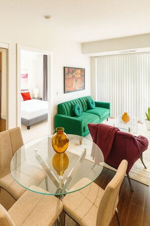 A modern hotel room with a glass dining table, a green sofa, a red armchair, and a bed with white bedding. - New · Monthly 1BR Condo by CN Tower & PATH (Toronto)