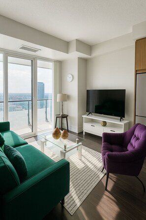 Una sala de estar moderna con un sofá verde, un sillón púrpura, una mesa de centro de vidrio, un televisor de pantalla plana sobre un soporte blanco, y un ventanal con vista a la ciudad.