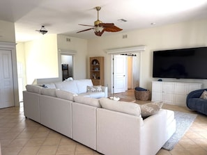 A living room with a sectional sofa, a flat-screen TV, a ceiling fan, and a large window. - Salty Serenity | Your Hernando Beach Escape (Hernando Beach)