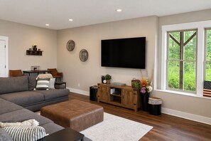 Ein modernes Wohnzimmer mit einem großen Flachbildfernseher, einem modularen Sofa, einem Hocker, einer Holzunterhaltungseinheit und einem großen Fenster mit Blick auf Grünflächen.