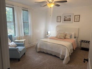 Ein Schlafzimmer mit einem Bett, einem Sessel, einem Nachttisch, einem Deckenventilator und einem Fenster mit Vorhängen.