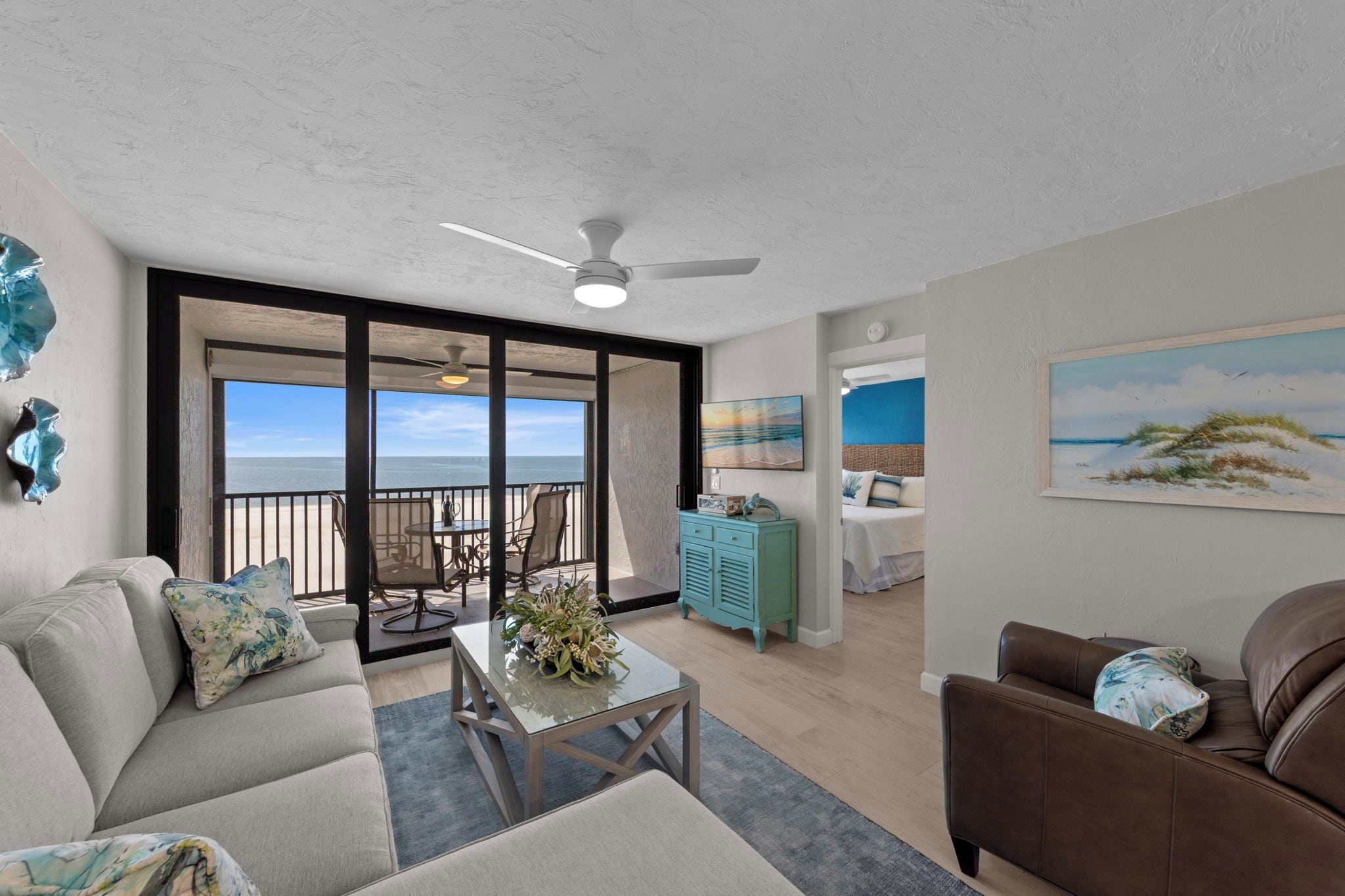 A spacious living area with a balcony overlooking the sea, a ceiling fan, and a dining table with chairs.