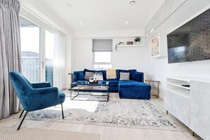 A modern living room with a blue sofa, a coffee table, and a blue armchair. There is a large window with curtains, a flat-screen TV mounted on the wall, and a small side table. - Modern London Apartment Near The O2, ExCeL & London City Airport - Sleeps 5 (London)