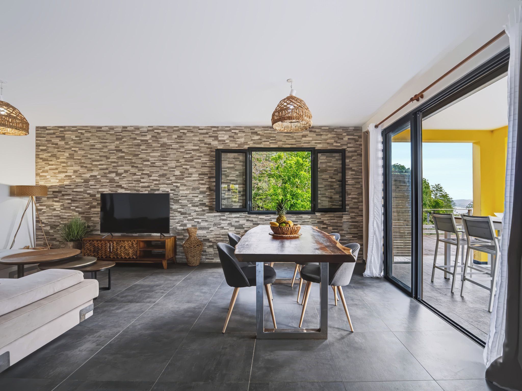 A modern living room with a flat-screen TV, a dining table with chairs, a large window, and a sliding glass door leading to an outdoor area.
