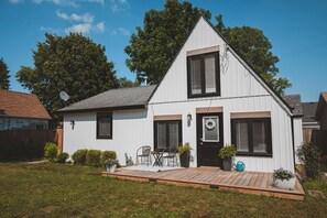 A white two-story house with a black roof, a front porch with a table and chairs, and a small garden area. - The Huron House - Lake Access (Brights Grove)