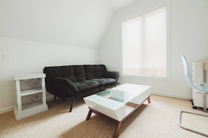 A room with a black sofa, a white coffee table, a white shelving unit, and a window with blinds. - The Huron House - Lake Access (Brights Grove)