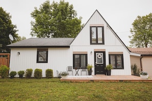 A white single-story house with a black door, two black-framed windows, a small porch with a chair, and a satellite dish. - The Huron House - Lake Access (Brights Grove)