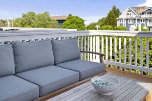 A grey outdoor sofa with cushions on a wooden deck, a white bowl with green items, and a white picket fence. - Rooftop Sunsets, EV Charger, Pool & Steps to Beach (New Buffalo)