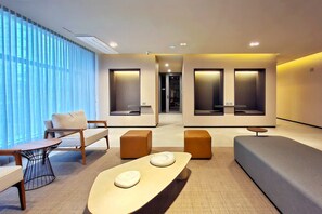 Ein modernes Hotelzimmer mit einer Couch, Sesseln, einem kleinen Tisch und einem großen Fenster mit Jalousien.