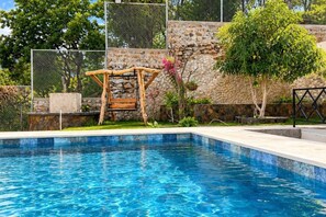 Una piscina con estructura de cubierta de madera, rodeada de un muro de piedra y vegetación.