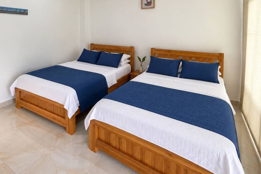 Dos camas de madera con ropa de cama blanca y azul, una planta pequeña en la mesita de noche y un cuadro en la pared.