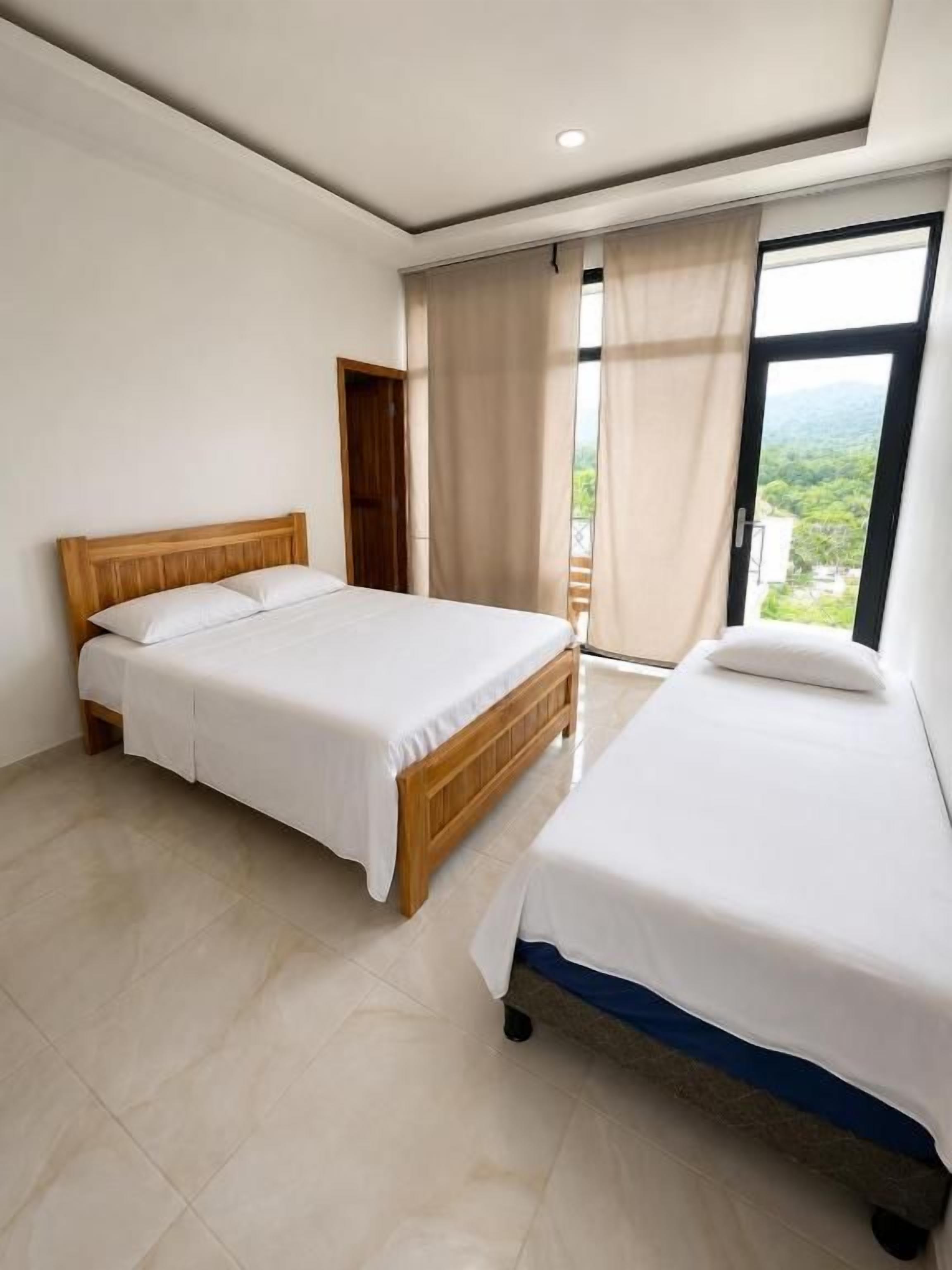 A bedroom with two beds, a wooden headboard, a large window with curtains, and a view of greenery outside.