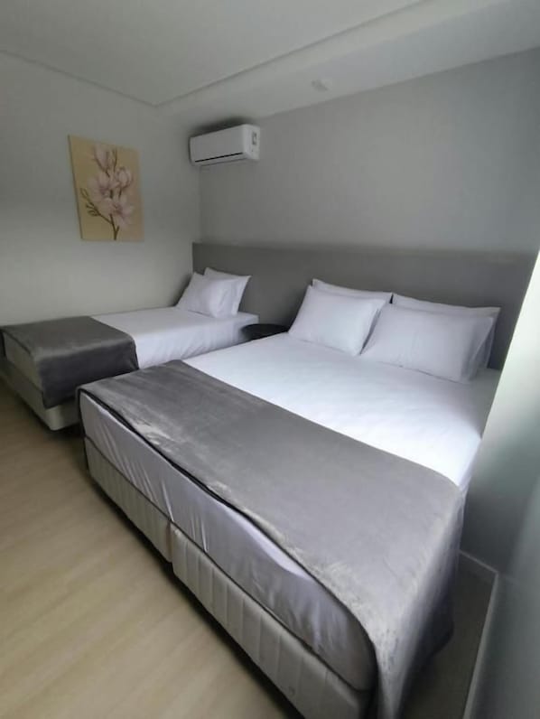 A hotel room with two beds, a grey sofa, a wooden floor, and a wall-mounted air conditioner.