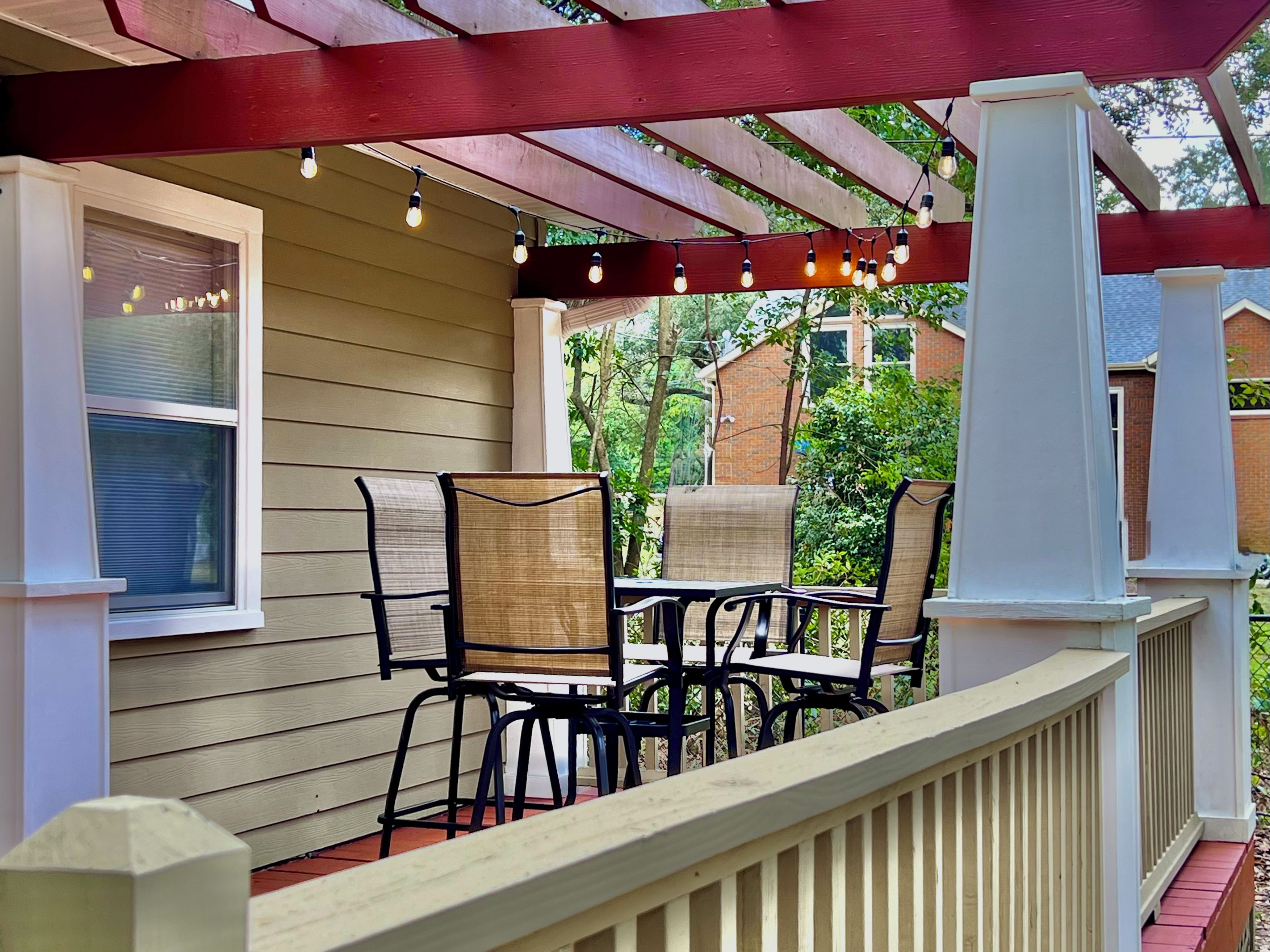 A covered outdoor seating area with a table and chairs, a string of lights, and a view of greenery.