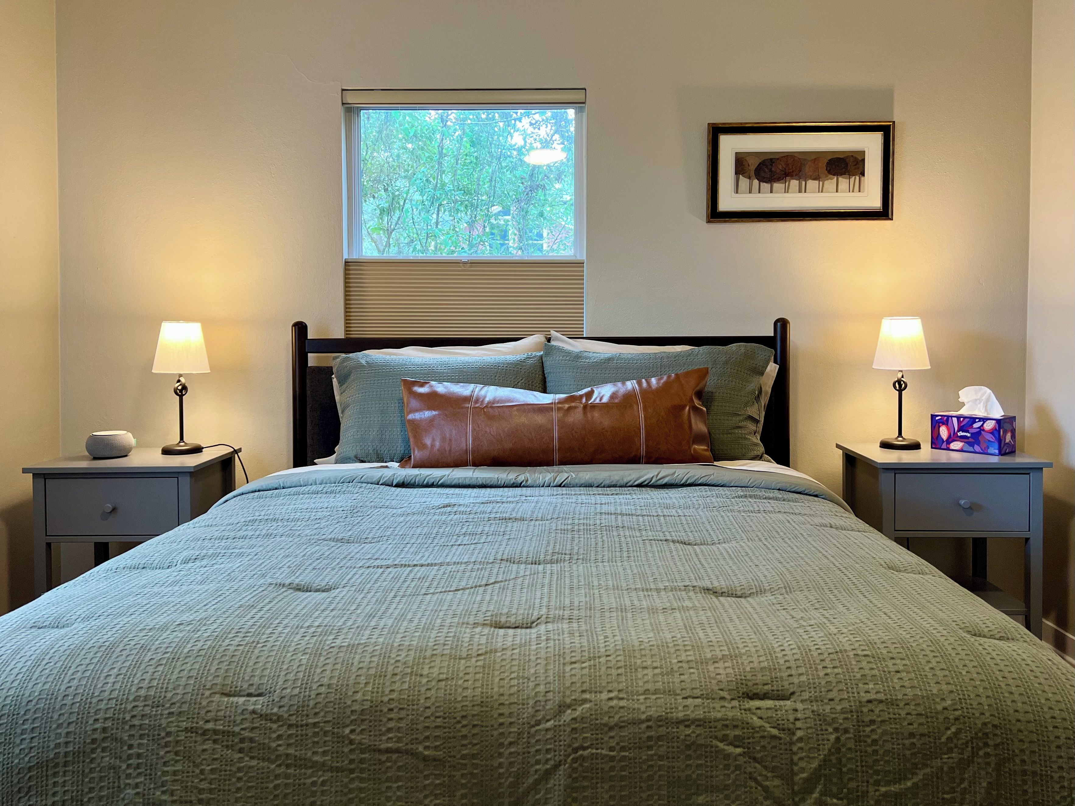 A bedroom with a bed, two bedside tables, a window with blinds, a framed picture on the wall, and a bedside lamp.