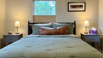 A bedroom with a bed, two bedside tables, a window with blinds, a framed picture on the wall, and a bedside lamp.