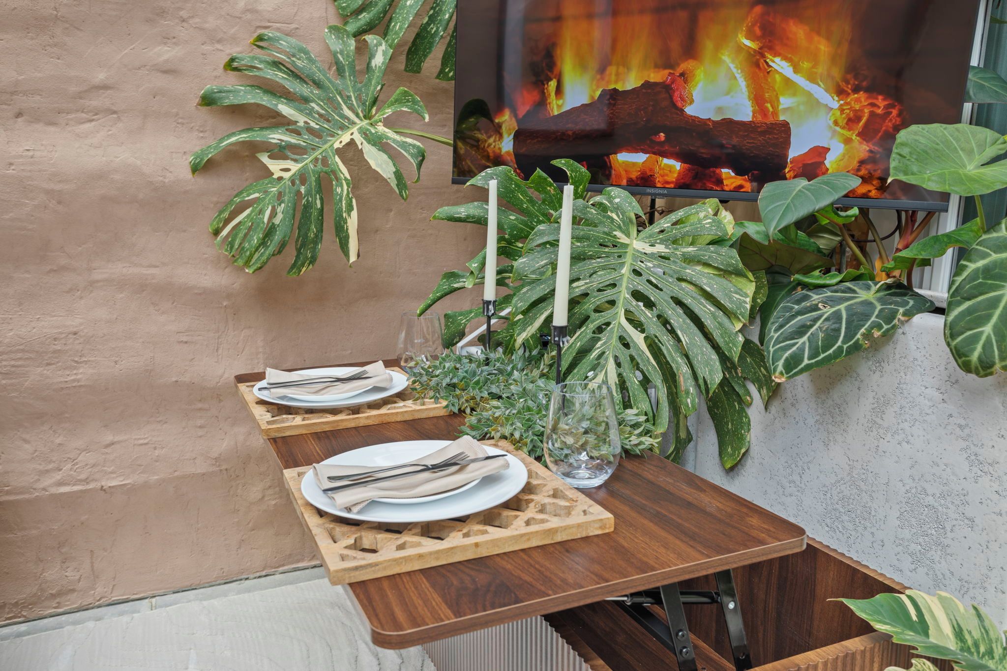 Un área de comedor al aire libre con mesas y sillas de madera, rodeada de grandes plantas verdes.