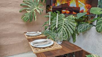Un área de comedor al aire libre con mesas y sillas de madera, rodeada de grandes plantas verdes.