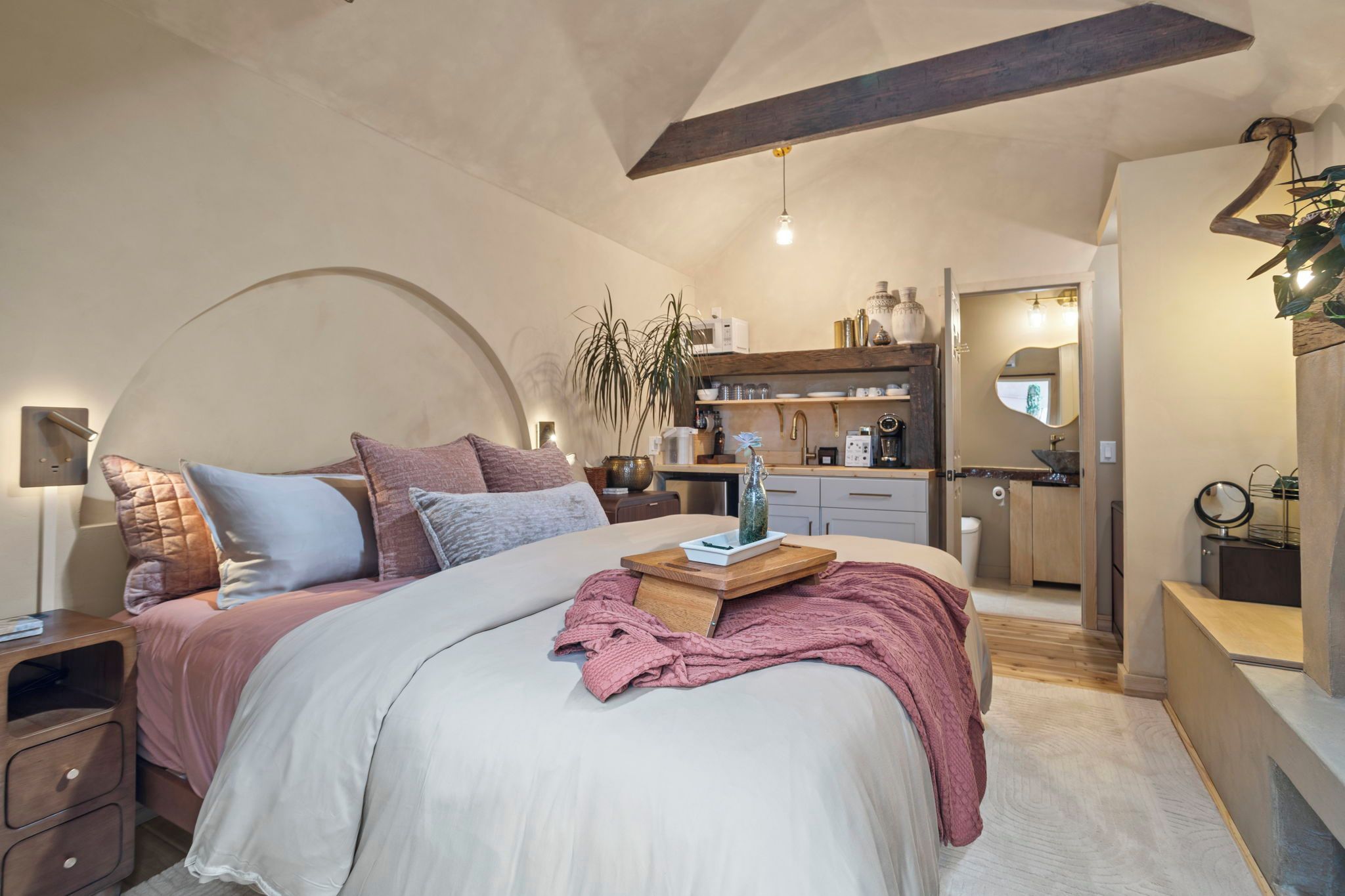Un dormitorio moderno con una cama grande, mesitas de noche, una lámpara de noche, una planta en maceta y vistas a la cocina contigua.