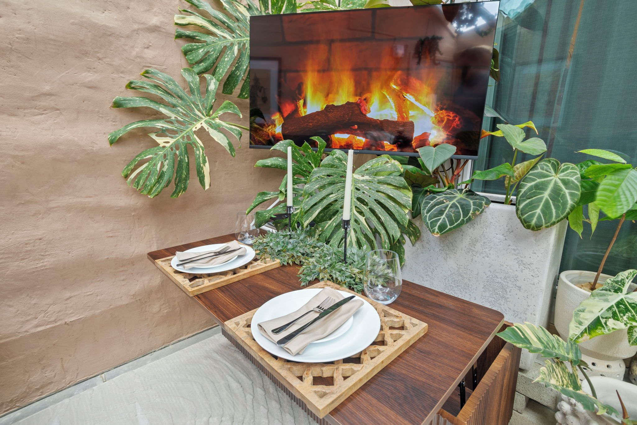 Une table à manger dressée avec des assiettes, des couverts et des bougies, entourée de grandes plantes vertes et d’un écran de télévision diffusant une image de feu de cheminée.