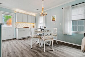 A kitchen with white cabinets, a dining table with chairs, a chandelier, and a window with curtains. - Cozy 2-bedroom house in serene North Myrtle Beach 1.5 blocks from beach (North Myrtle Beach)