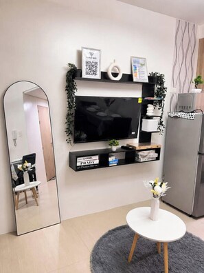 A modern living room with a flat-screen TV mounted on the wall, a small table with a vase, and a refrigerator. - STAYCATION SUITES PH 2 (Pasay City)