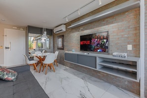 Een moderne woonkamer met een bakstenen muur, een flatscreen televisie die boven een console hangt, en een eethoek met witte stoelen en een glazen tafel.