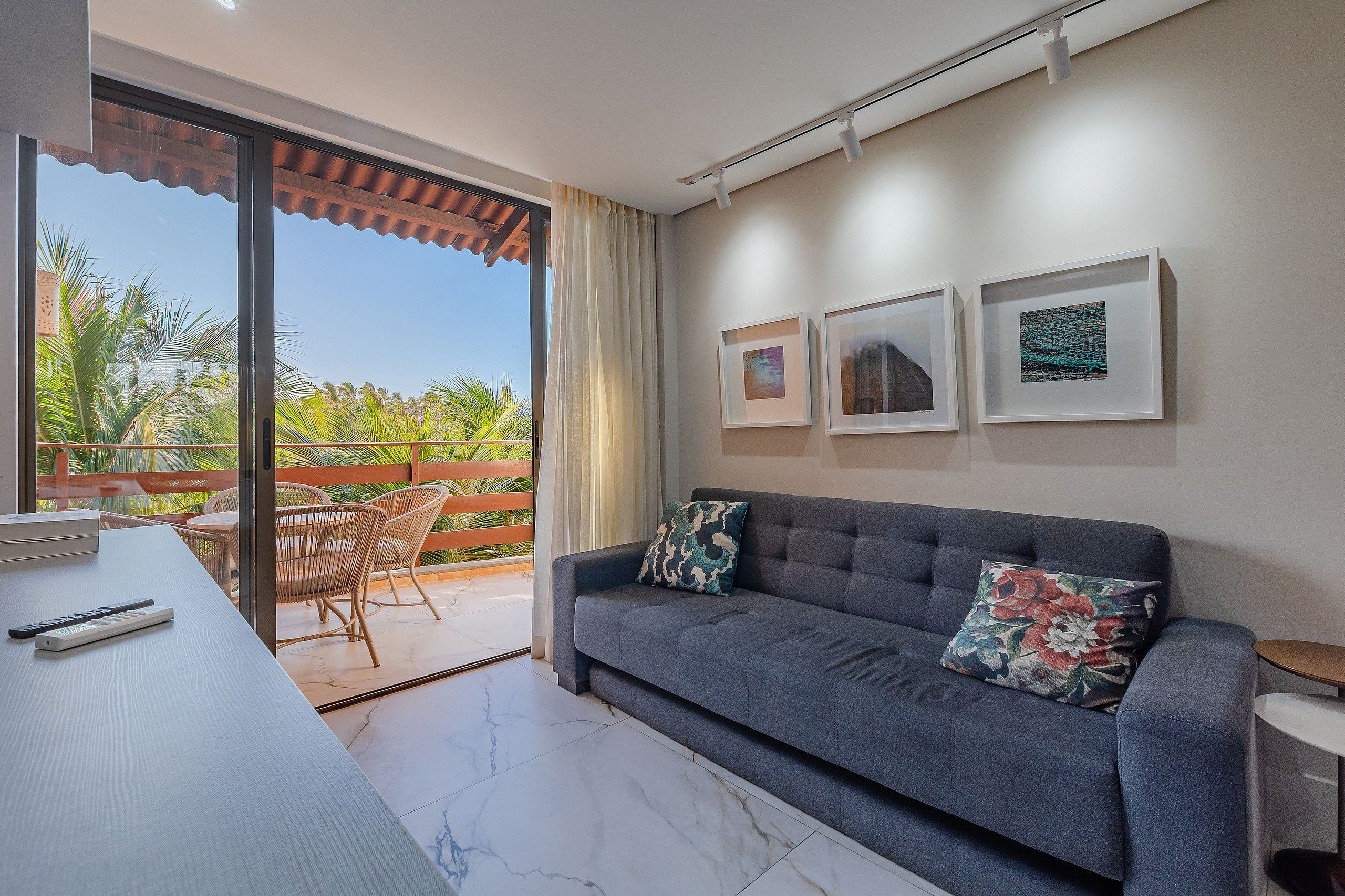 Sala de estar com sofá, mesa e varanda com cadeiras. Três quadros emoldurados na parede.
