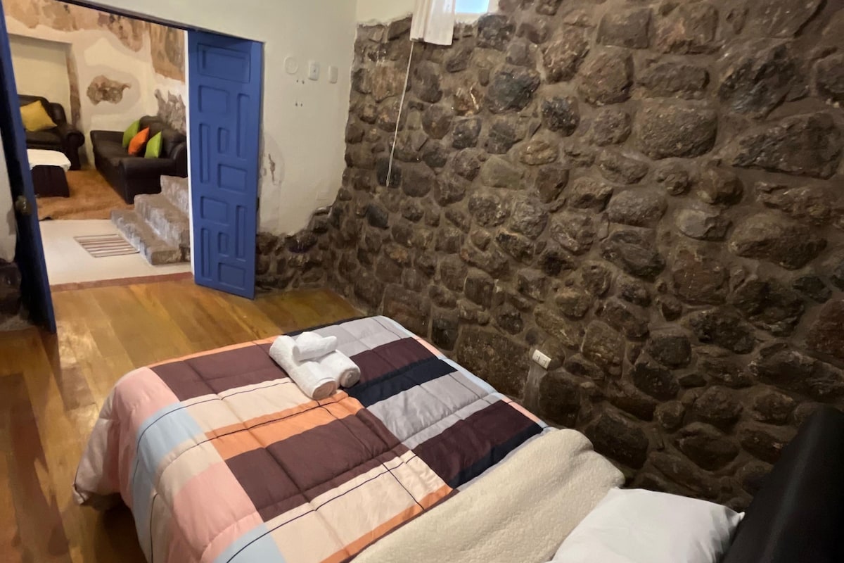 A bedroom with a stone wall, a bed with a quilt, a wooden floor, and a door leading to another room.