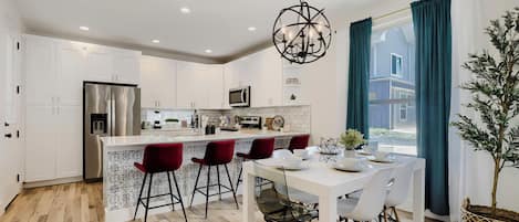 A modern kitchen with white cabinetry, a stainless steel refrigerator, a central island with bar stools, and a hanging light fixture.