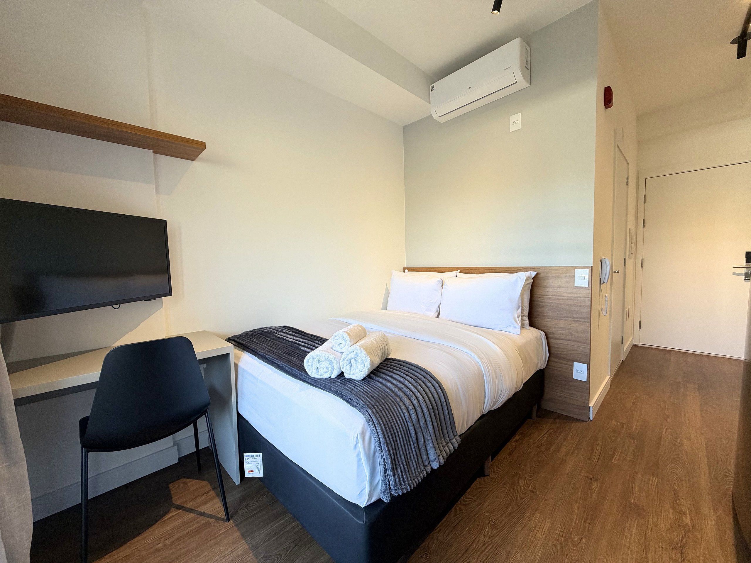 A hotel room with a bed, a desk, a chair, an air conditioning unit, and a door.