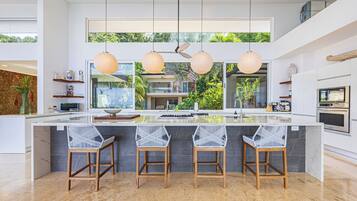 Une cuisine moderne dotée d’un grand îlot central, de luminaires suspendus et offrant une vue sur l’extérieur.