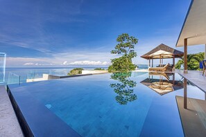 A modern infinity pool with a clear view of the ocean, a wooden lounge area with umbrellas, and a glass railing. - Casa Ombak 7-Bedroom Luxury Villa with Spa & Ocean View (Puntarenas Province)