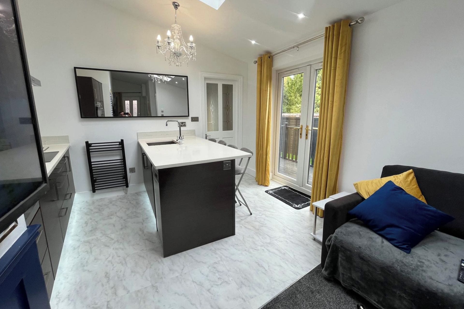 A modern kitchen with a central island, a dining area, and a living room with a sofa and cushions.