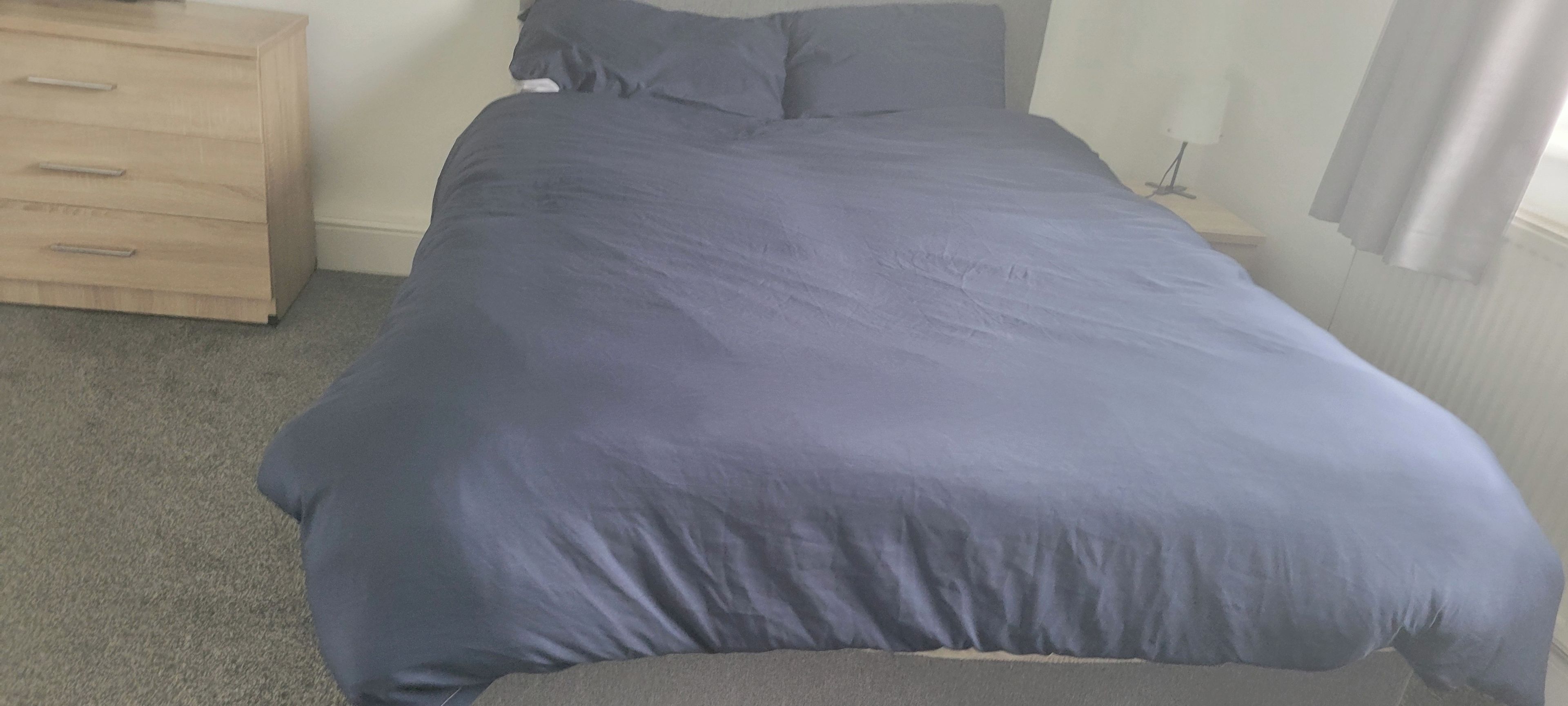 Una cama con un colchón gris y una manta azul en una habitación con un aparador de madera y una mesita de noche.