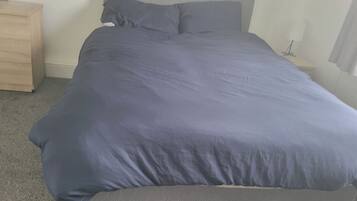 A bed with a grey mattress and a blue blanket in a room with a wooden dresser and a nightstand.