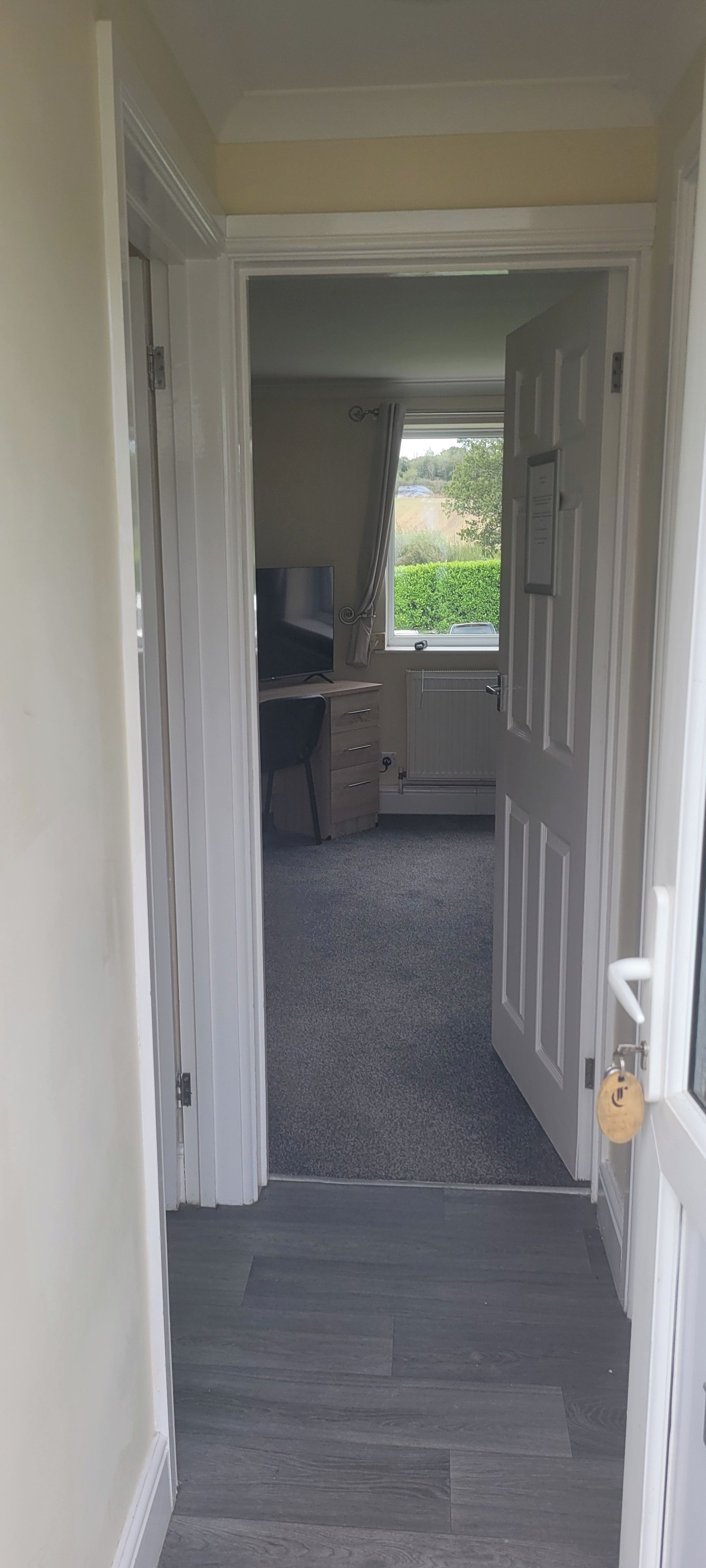 Un couloir mène à une pièce où se trouvent un bureau et une chaise, un radiateur et une fenêtre donnant sur un paysage verdoyant.