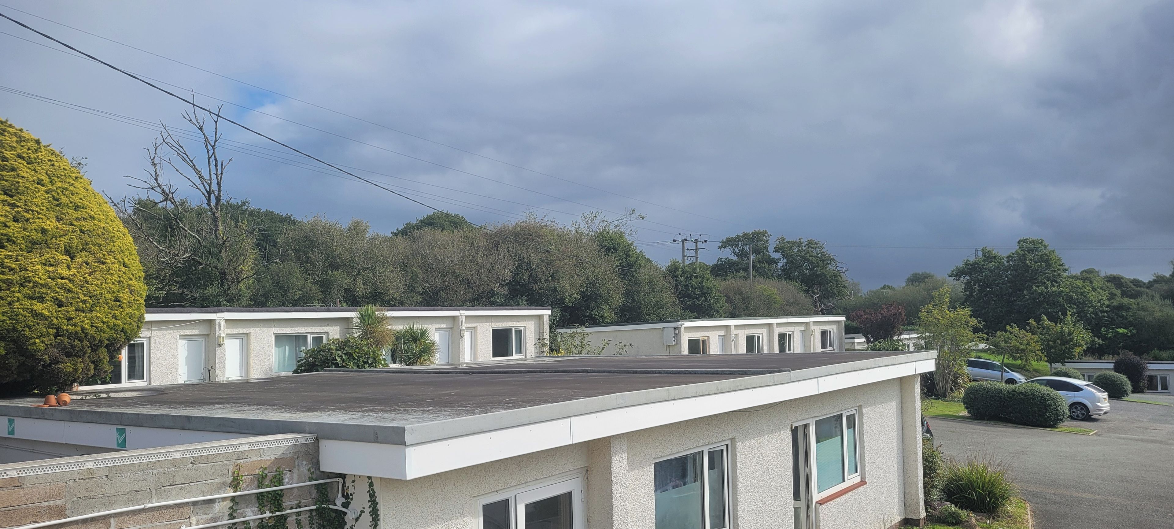 Una hilera de edificios de una planta con techos planos, rodeados de árboles y coches aparcados.