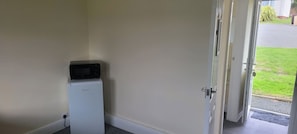 Een kleine kamer met een witte koelkast, een magnetron erboven en een deur naar een grasrijk buitengebied.