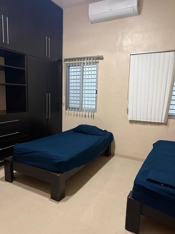 A room with two single beds, a wardrobe, a window with bars, and an air conditioning unit. - Departamentos Gonzalo Guerrero (Mérida)