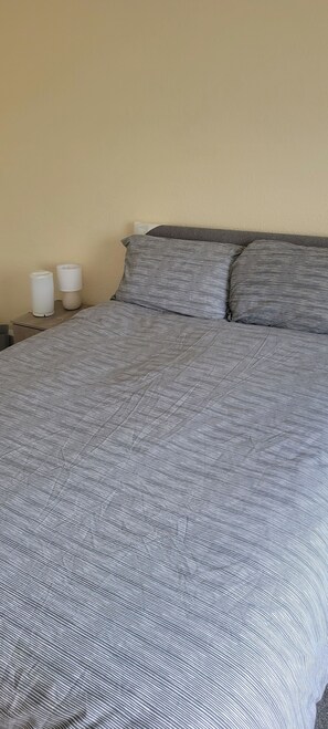 Ein Bett mit gestreifter Bettdecke und zwei Kissen, ein Nachttisch mit Lampe sowie eine Wand mit strukturierter Oberfläche.