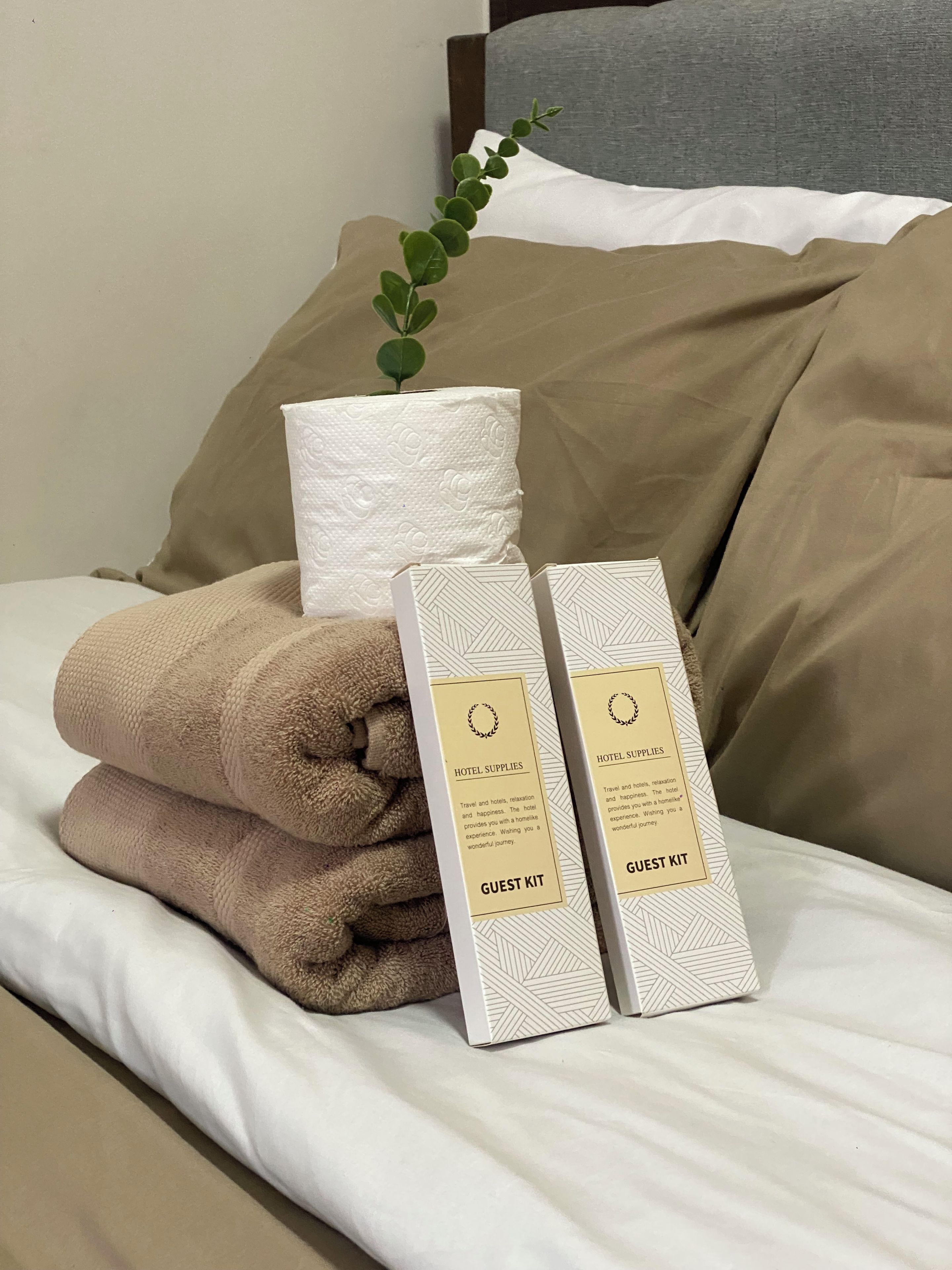 A neatly made bed with a beige pillow, a rolled-up towel, and two guest kits on a white bedspread.