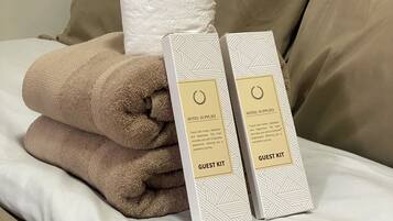 A neatly made bed with a beige pillow, a rolled-up towel, and two guest kits on a white bedspread.