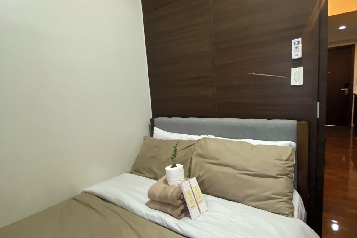 A hotel room with a bed, a nightstand, a wall-mounted thermostat, and a door leading to another room.