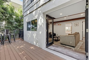Ein modernes Haus mit einer Holzterrasse, einer Glastür, die in ein Wohnzimmer mit Sofa und Couchtisch führt, und einem kleinen Essbereich.