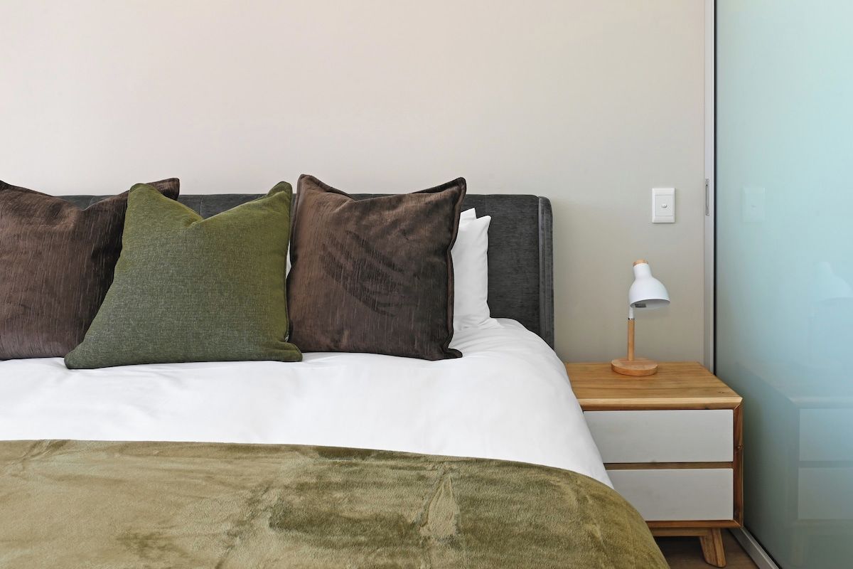 A bed with a white duvet, a green throw blanket, and multiple pillows. There is a wooden nightstand with a white lamp beside the bed.