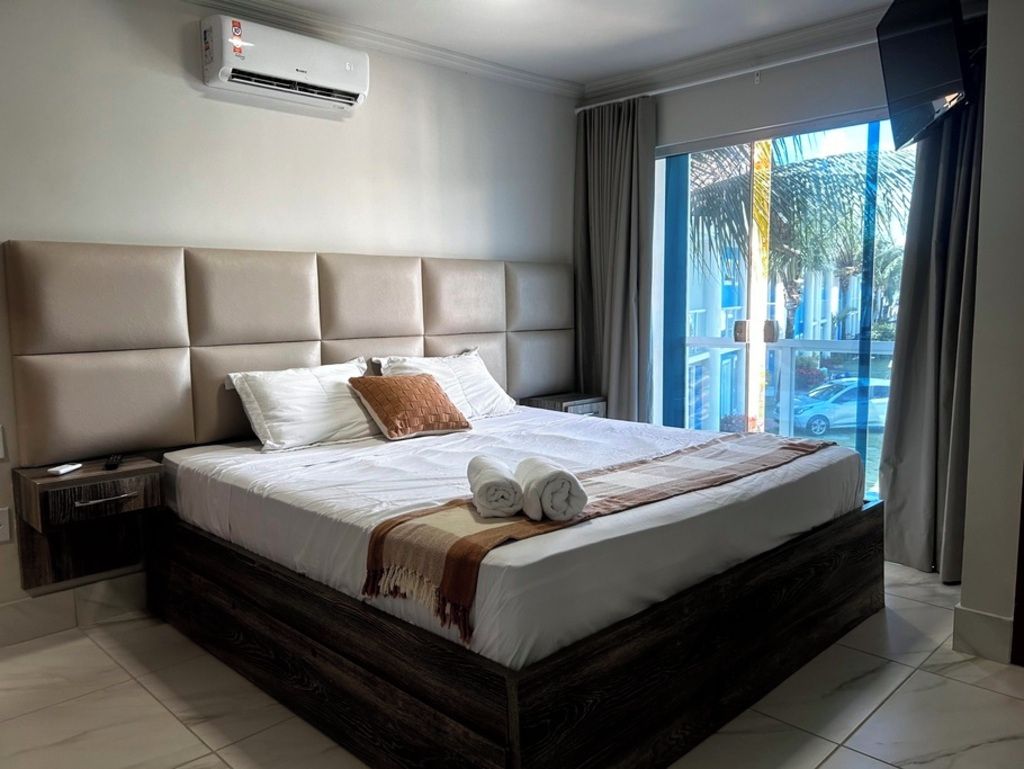 A hotel room with a large bed, a nightstand, an air conditioning unit, and a view of the outside.