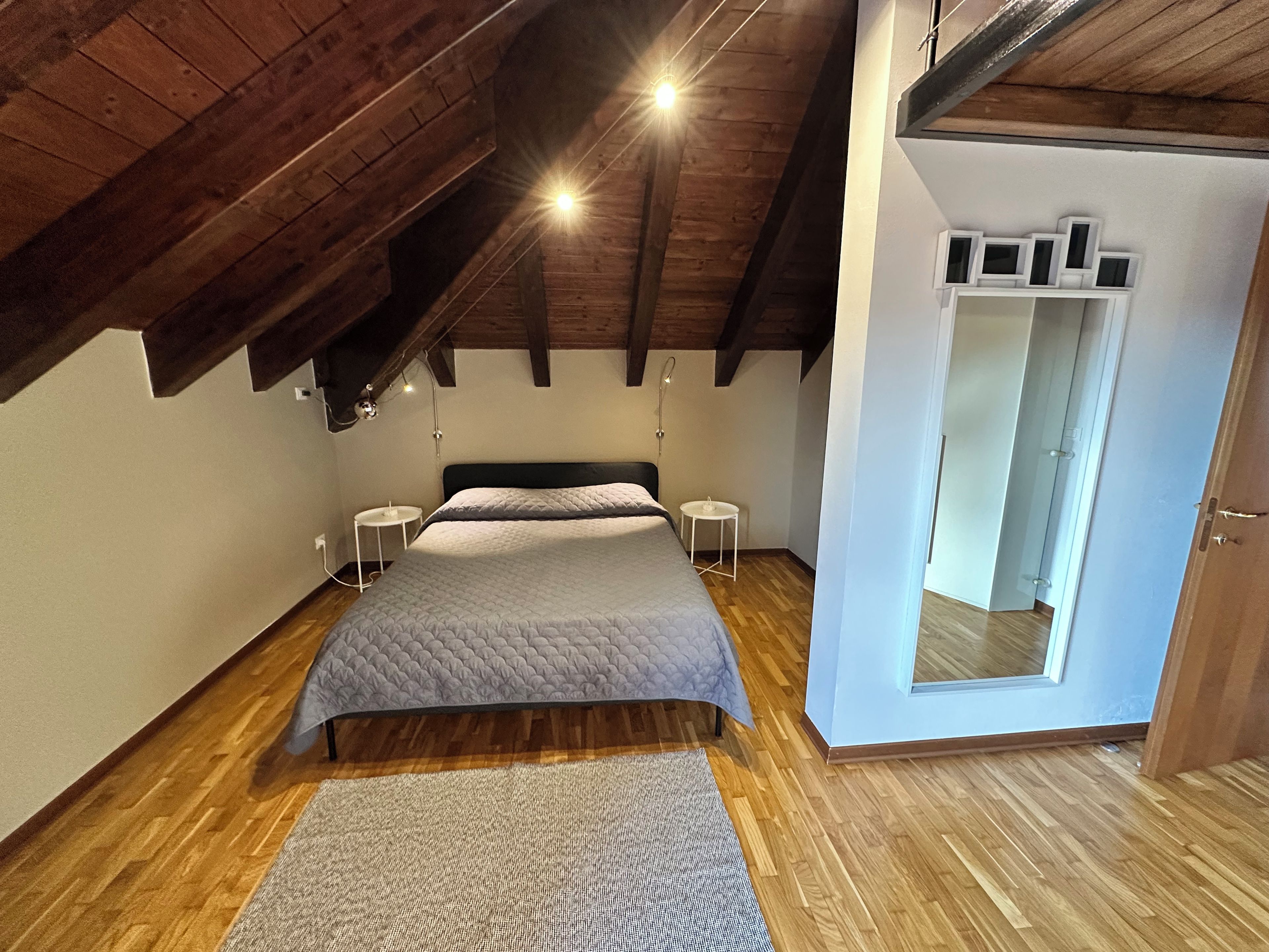Ein Schlafzimmer mit Holzboden, einem Bett mit grauer Decke, zwei Nachttischen und einem Spiegel an der Wand.